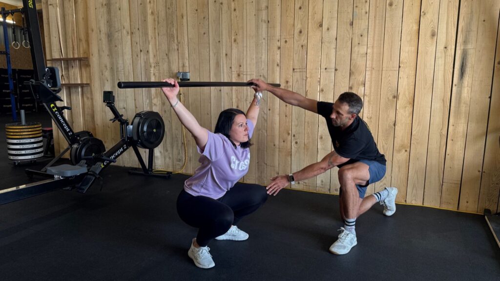 Un coach corrige la position d'une adhérente en squat, bâton au-dessus de la tête, à la salle CrossFit Hormesis de Bourg-Blanc